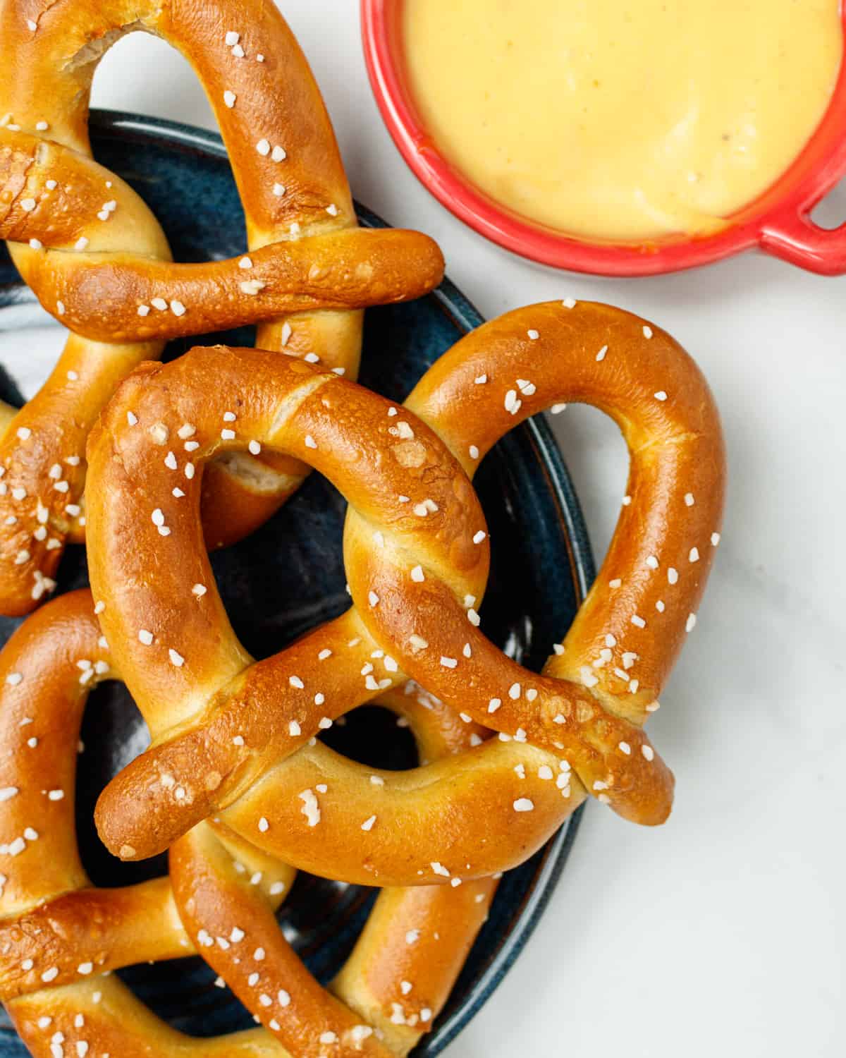 Three soft pretzels on a dark blue plate sitting on a white background. A small red bowl of mustard is in the top right of the photo.