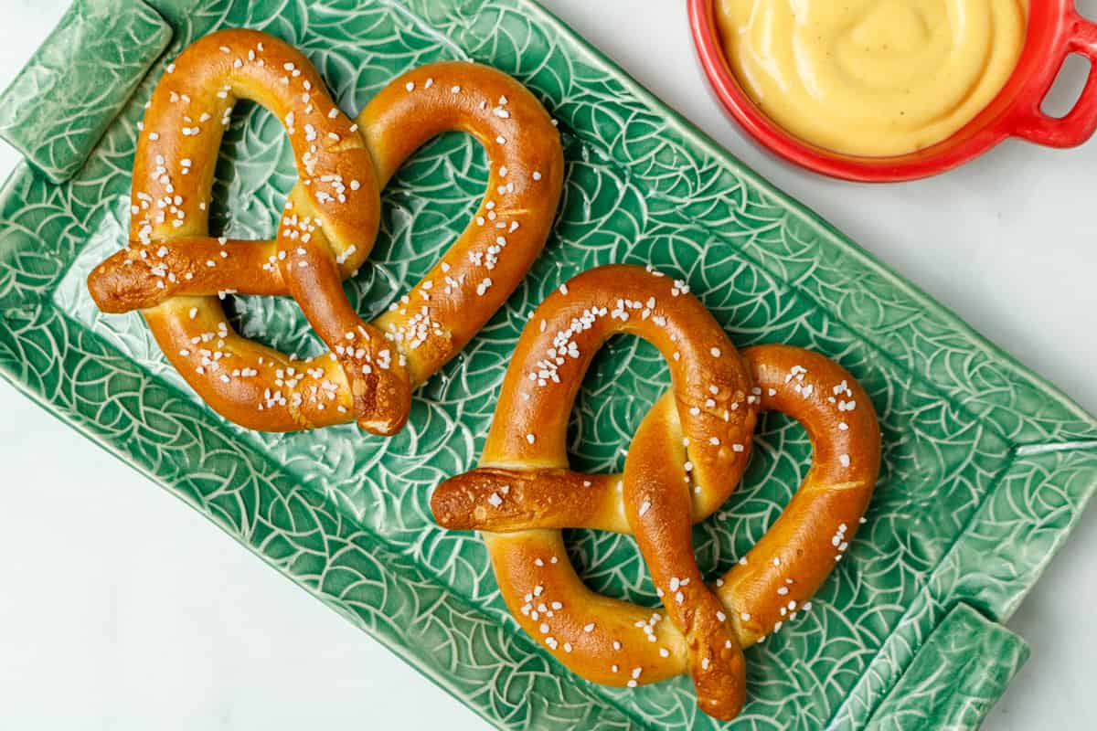 Two soft pretzels on a green platter. In the corner is a red bowl with mustard.